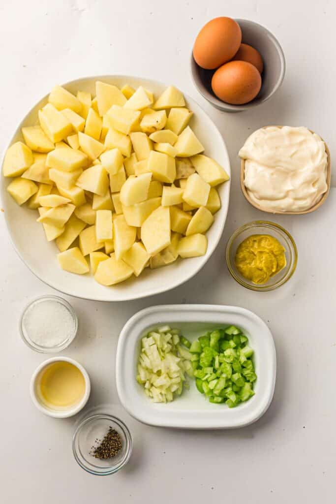 A white surface with bowls containing chopped potatoes, chopped celery, chopped onion, mayonnaise, mustard, two whole eggs, salt, pepper, and vinegar, arranged neatly for a potato salad recipe.