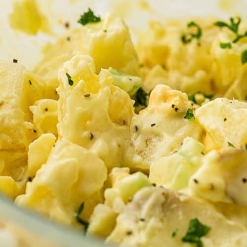 Close-up of creamy potato salad with chunks of potato, finely chopped celery, black pepper, and parsley sprinkled on top. The mixture appears to be coated in a mayonnaise-based dressing.