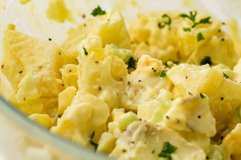 Close-up of creamy potato salad with chunks of potato, finely chopped celery, black pepper, and parsley sprinkled on top. The mixture appears to be coated in a mayonnaise-based dressing.