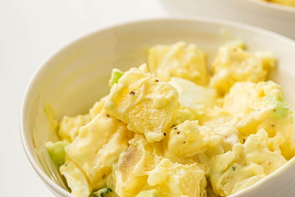 A close-up of a bowl filled with creamy potato salad, featuring chunks of potatoes mixed with mayonnaise, celery, and a sprinkle of black pepper.