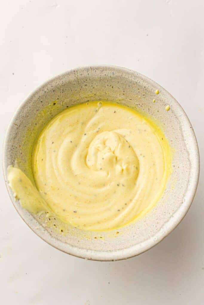 A ceramic bowl filled with creamy, yellow mayonnaise, swirled smoothly with specks of seasoning, sits on a white surface. Some mayonnaise is spread on the rim of the bowl.