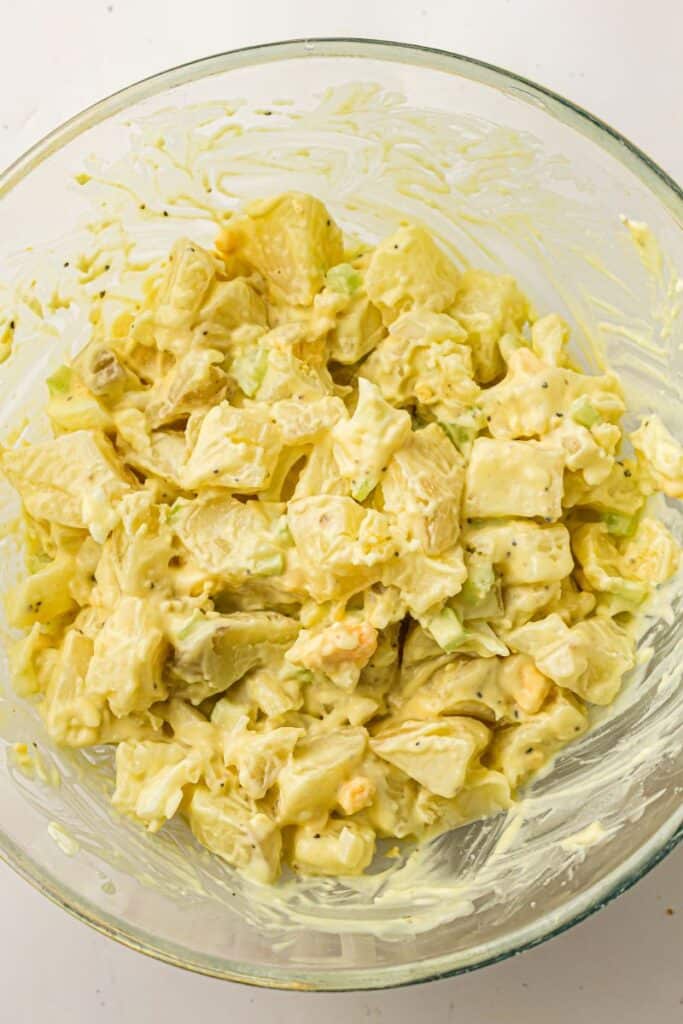 A glass bowl filled with creamy potato salad, featuring diced potatoes, chopped celery, and pieces of egg, all coated in a yellowish mayonnaise-based dressing.