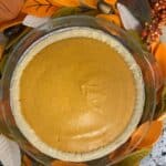 A pumpkin pie with a smooth orange filling sits in a pie dish, surrounded by autumn-themed decorations with orange, yellow, and green felt leaves, and small faux acorns and berries.