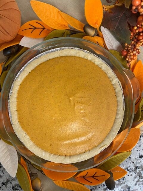 A pumpkin pie with a smooth orange filling sits in a pie dish, surrounded by autumn-themed decorations with orange, yellow, and green felt leaves, and small faux acorns and berries.