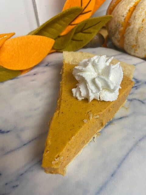 A slice of pumpkin pie with a dollop of whipped cream on top sits on a marble surface, with autumn-themed felt leaves and a small decorative pumpkin in the background.