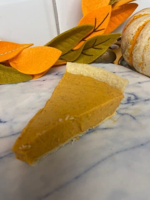 A slice of pumpkin pie with a light, flaky crust sits on a marble countertop. In the background, there is a decorative felt autumn leaf garland and part of a small pumpkin.
