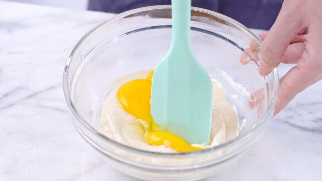 A hand uses a light blue spatula to mix egg yolks into a creamy batter in a clear glass bowl on a white marble surface.