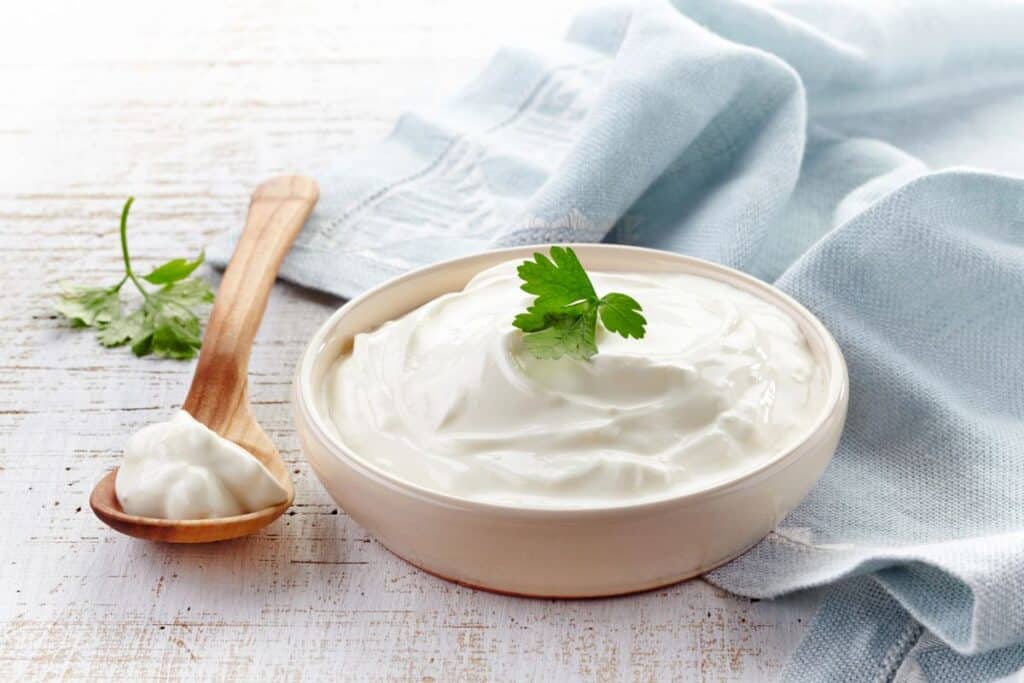 A bowl of creamy yogurt topped with a parsley leaf sits on a white wooden surface next to a blue cloth. A wooden spoon with a dollop of yogurt rests beside the bowl.