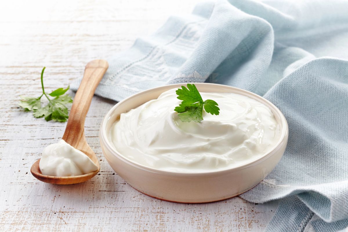 A bowl of creamy yogurt topped with a parsley leaf sits on a white wooden surface next to a blue cloth. A wooden spoon with a dollop of yogurt rests beside the bowl.