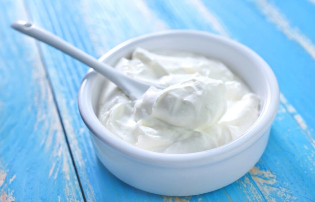 A white bowl filled with creamy yogurt sits on a blue wooden surface, with a white spoon resting inside the bowl.