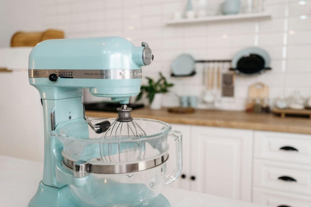 A light blue stand mixer with a glass bowl sits on a kitchen counter. The background features white cabinets, a wooden countertop, and various kitchen utensils and dishes against a white tiled wall.
