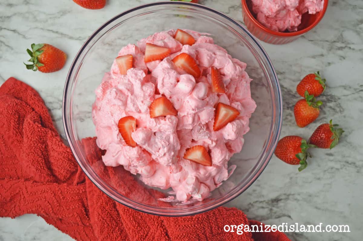 A glass bowl filled with creamy pink strawberry fluff dessert topped with fresh strawberry pieces, next to a red towel, loose strawberries, and another small bowl of fluff on a marble surface.