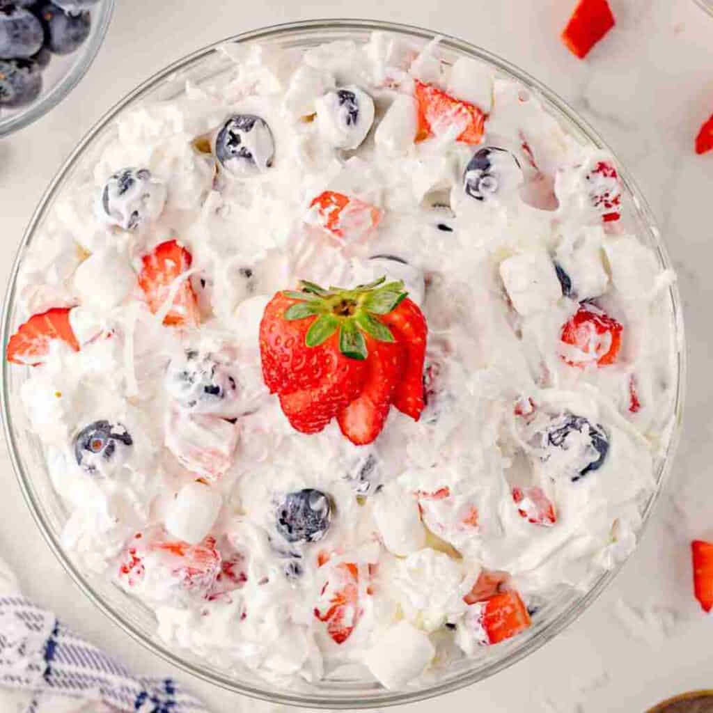 A glass bowl filled with creamy fruit salad, containing strawberries, blueberries, mini marshmallows, and shredded coconut, garnished with sliced strawberries on top.