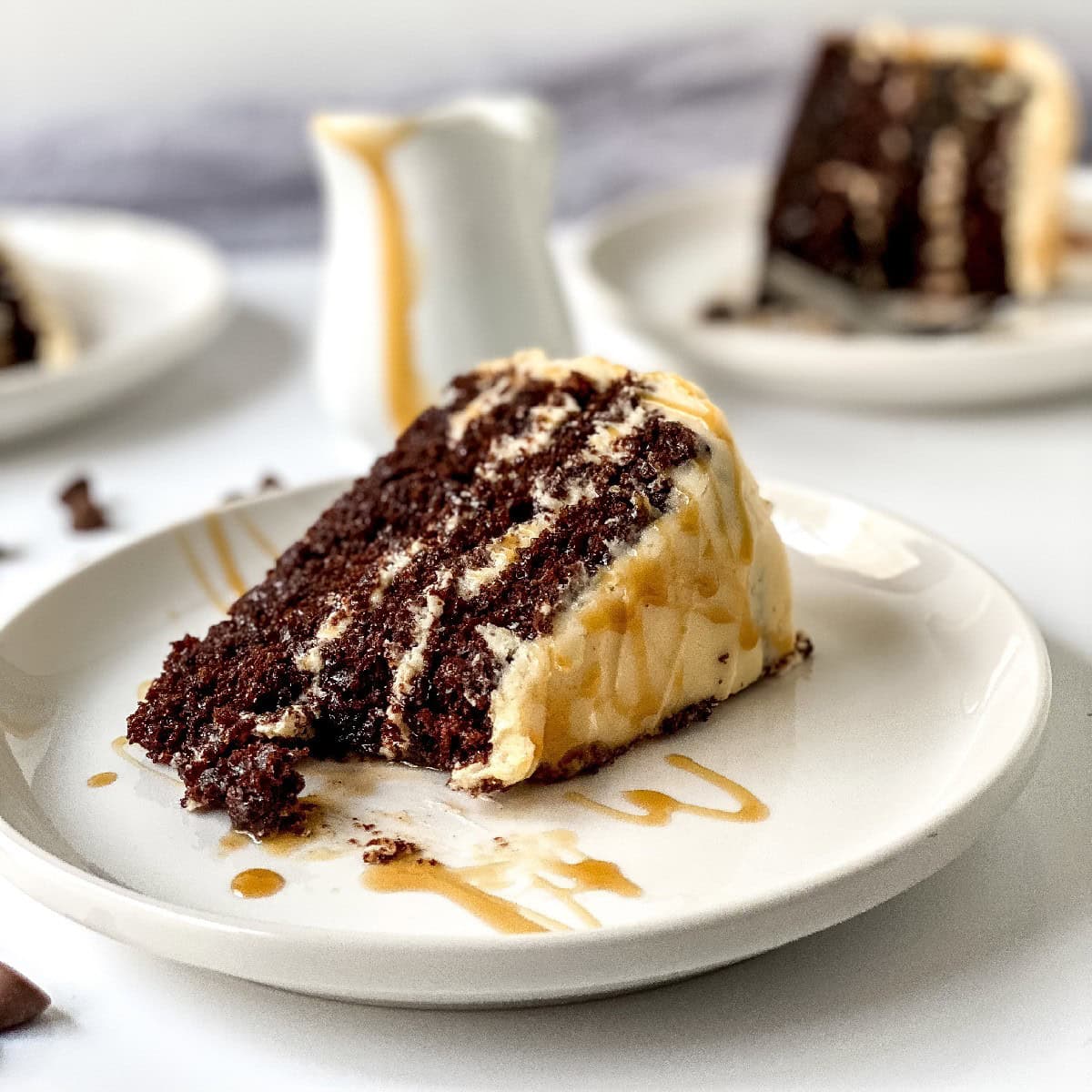 A slice of chocolate layer cake with cream filling and caramel drizzle sits on a white plate, with more cake slices and a small pitcher of caramel sauce in the background.