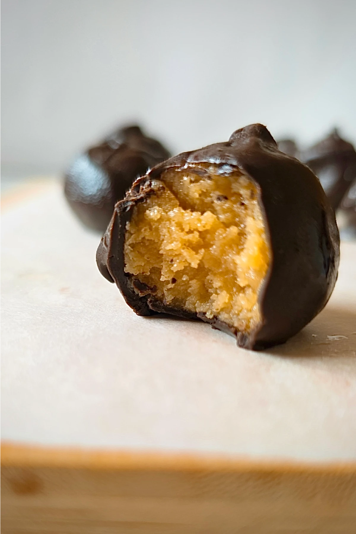 A close-up of a chocolate truffle with a bite taken out, revealing a creamy, golden filling inside. Other whole truffles are blurred in the background on a light-colored surface.
