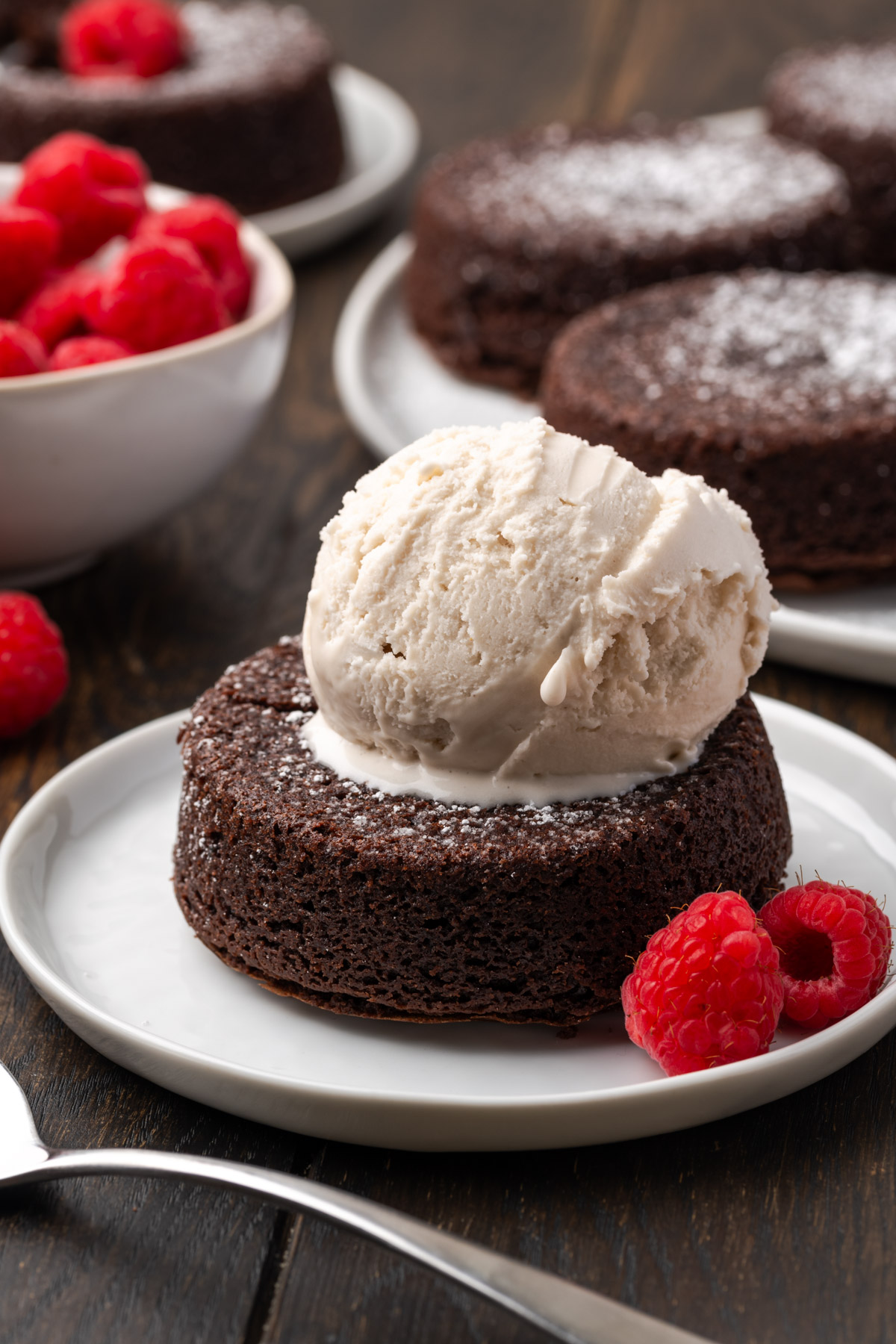 A plate with a chocolate lava cake topped with a scoop of vanilla ice cream and garnished with fresh raspberries; more lava cakes and a bowl of raspberries are in the background.