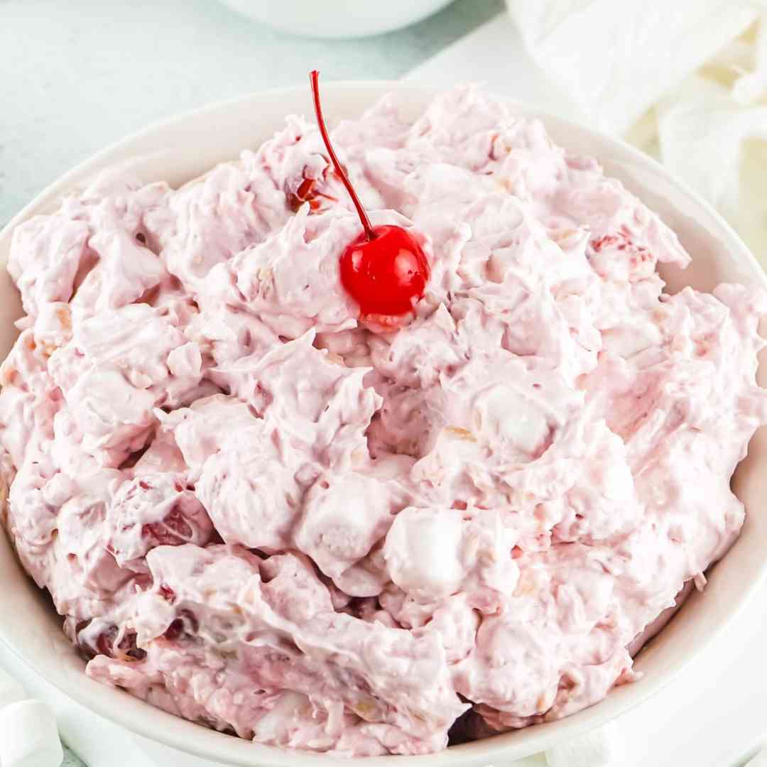 A white bowl filled with pink, fluffy ambrosia salad topped with a single red cherry. The salad contains visible chunks of fruit and marshmallows.