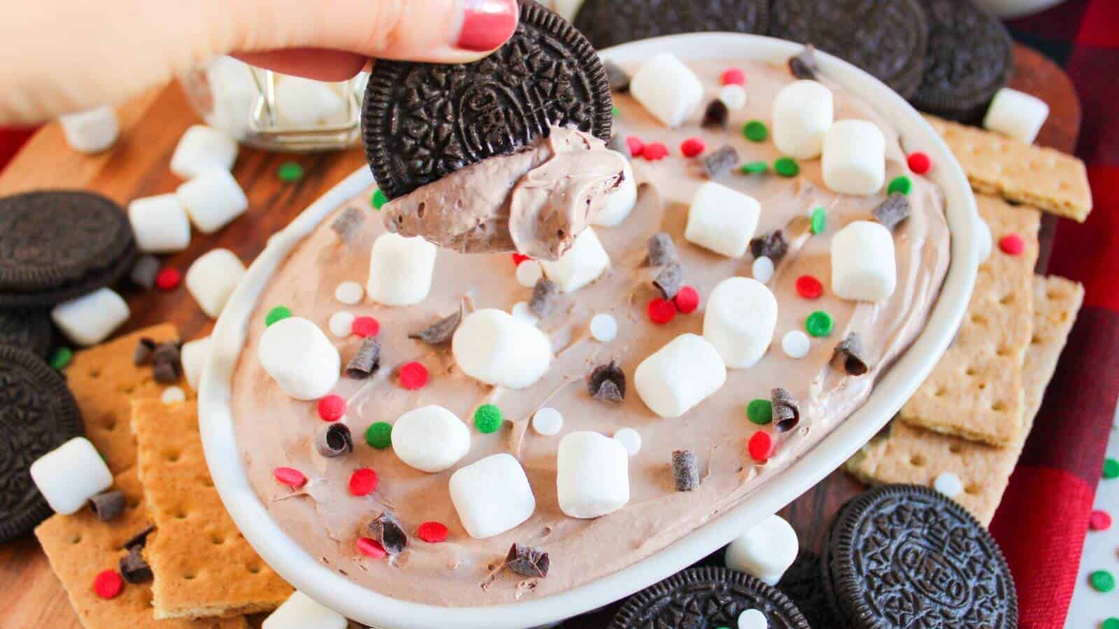 A hand dips a chocolate sandwich cookie into a creamy dessert topped with mini marshmallows, chocolate chips, and red and green sprinkles; graham crackers and cookies surround the dish.