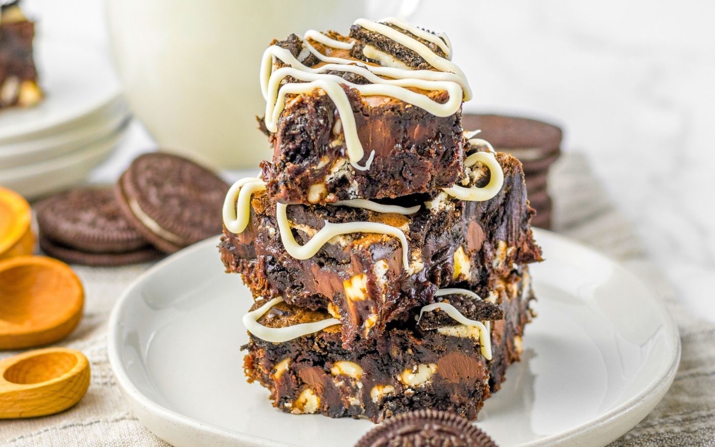 Three stacked chocolate brownies with chunks of cookies and white chocolate drizzle on top, served on a white plate. Whole chocolate sandwich cookies and wooden spoons are blurred in the background.