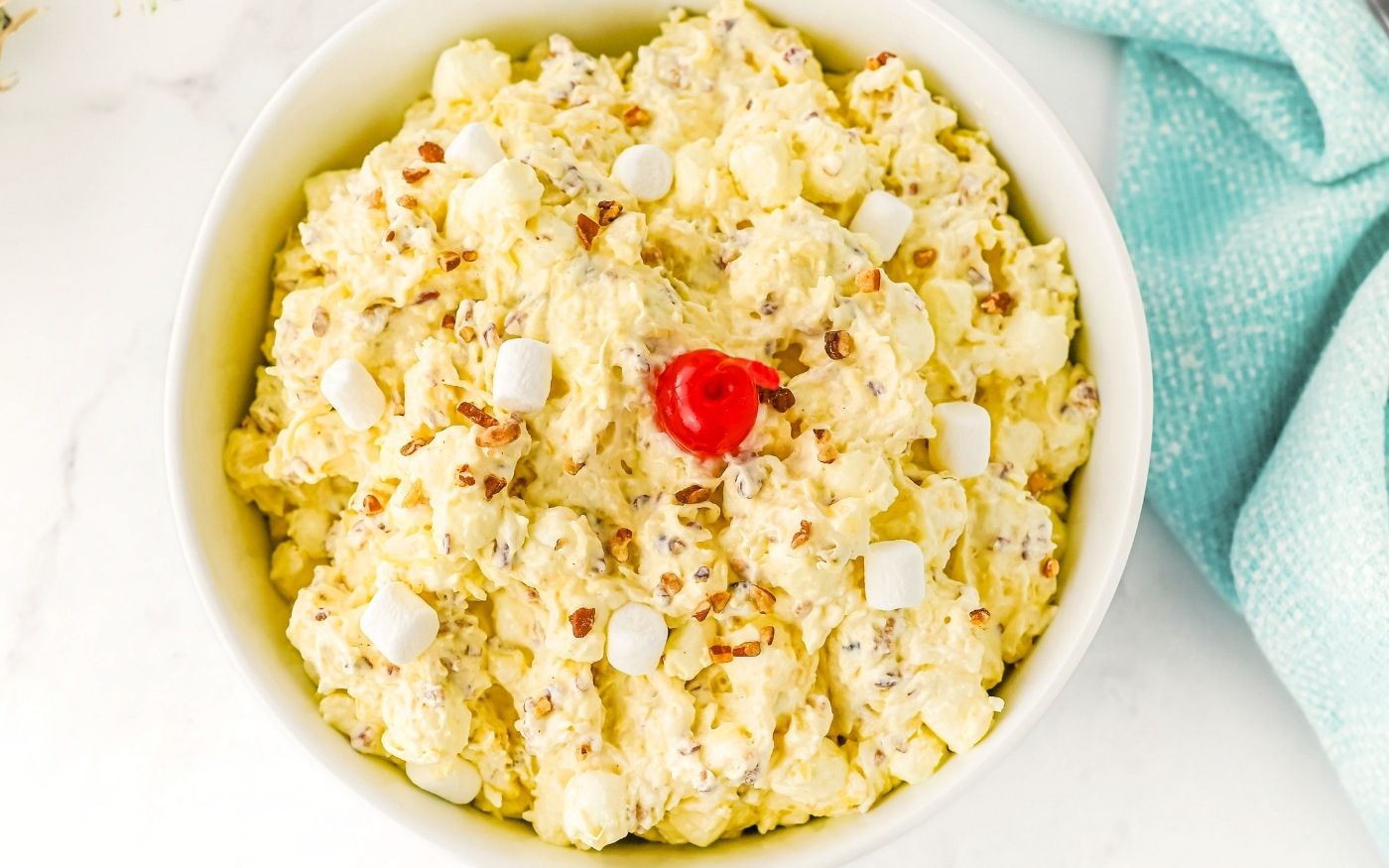 A bowl of creamy ambrosia salad topped with a single red cherry in the center, small white marshmallows, and sprinkled with chopped nuts. The bowl sits on a white surface next to a light blue cloth.