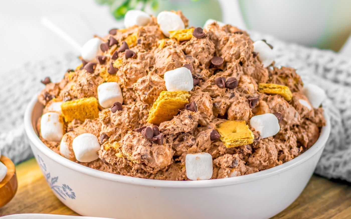 A large white bowl filled with a fluffy chocolate mixture, topped with mini marshmallows, chocolate chips, and graham cracker pieces, resembling a smores dessert salad.