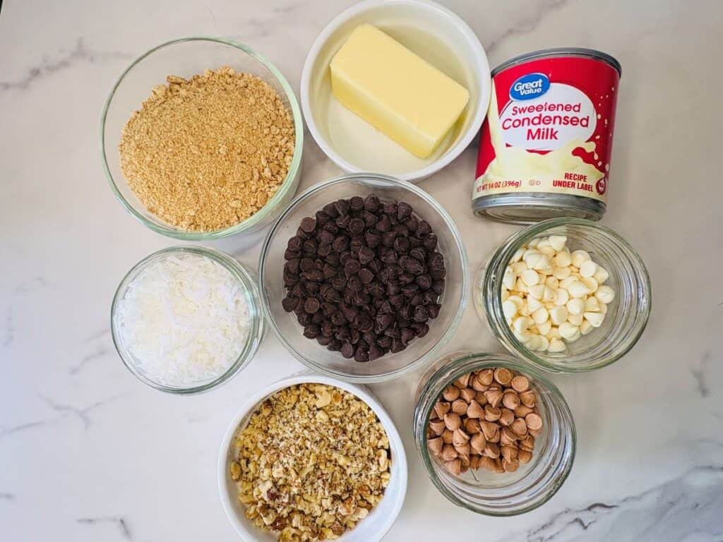 Eight baking ingredients are arranged on a white surface: graham cracker crumbs, shredded coconut, chopped walnuts, butterscotch chips, dark chocolate chips, white chocolate chips, a stick of butter, and a can of sweetened condensed milk.