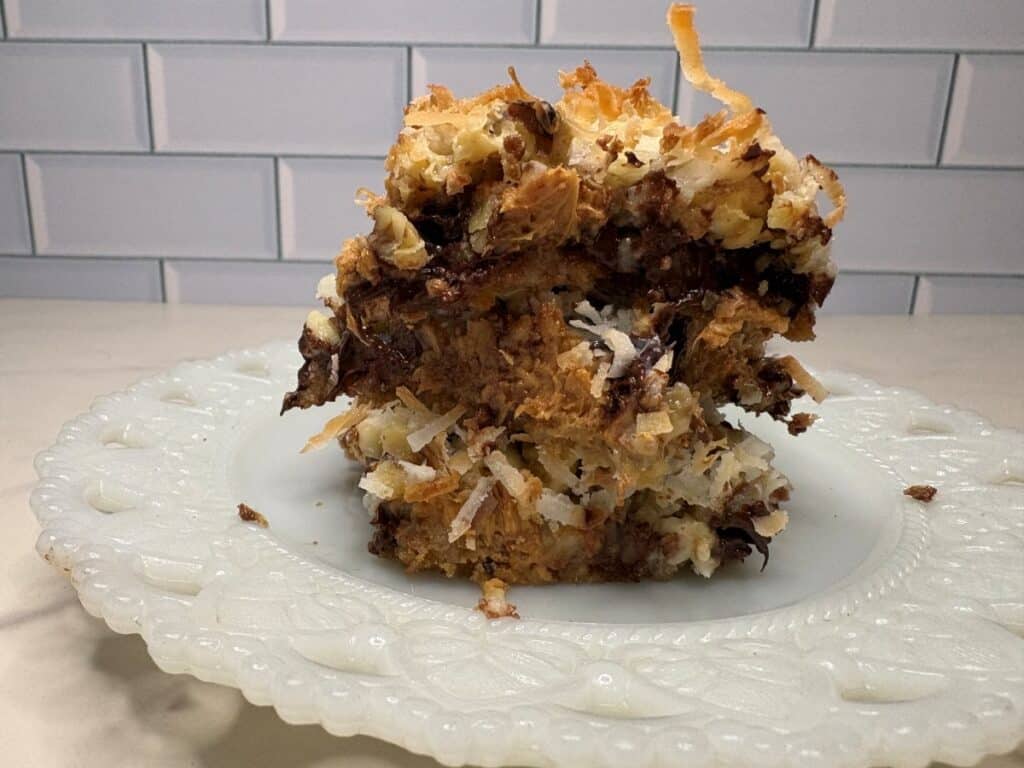 A close-up of a thick, square piece of dessert with layers of toasted coconut, chocolate, and a crumbly base, served on a decorative white plate in front of a white subway tile background.