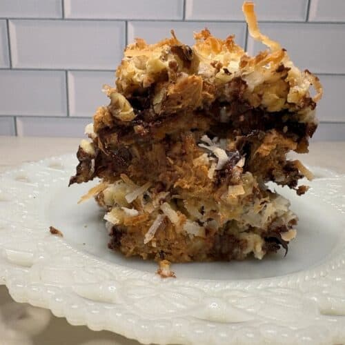 A close-up of a thick, square piece of dessert with layers of toasted coconut, chocolate, and a crumbly base, served on a decorative white plate in front of a white subway tile background.
