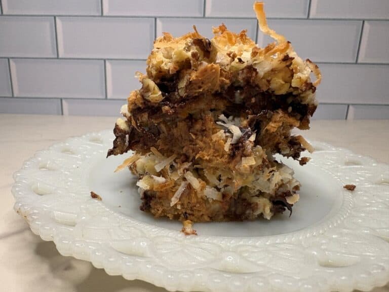A close-up of a thick, square piece of dessert with layers of toasted coconut, chocolate, and a crumbly base, served on a decorative white plate in front of a white subway tile background.