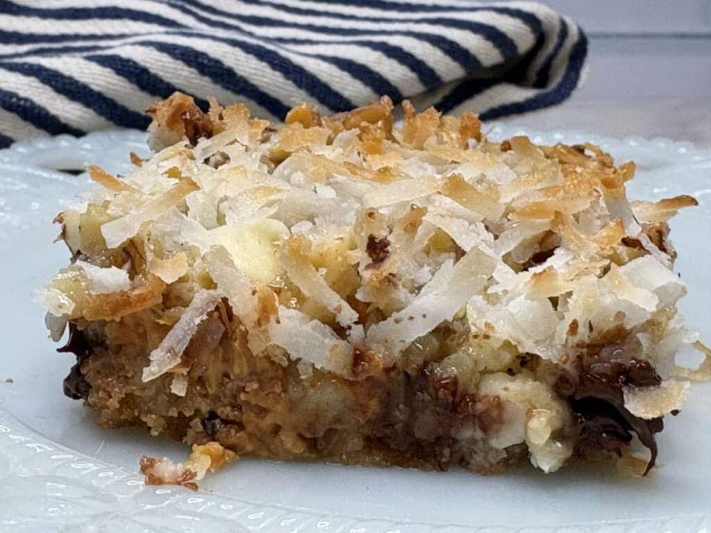 A close-up of a dessert bar topped with shredded coconut on a white plate, with a striped cloth in the background. The bar appears to have layers of chocolate, coconut, and a crumbly base.
