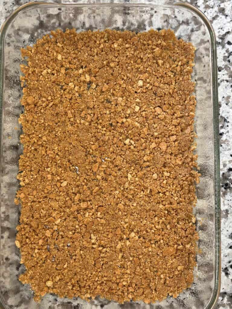 A rectangular glass dish filled with an even layer of golden-brown crumb mixture, likely a graham cracker or cookie crust, sits on a speckled granite countertop.