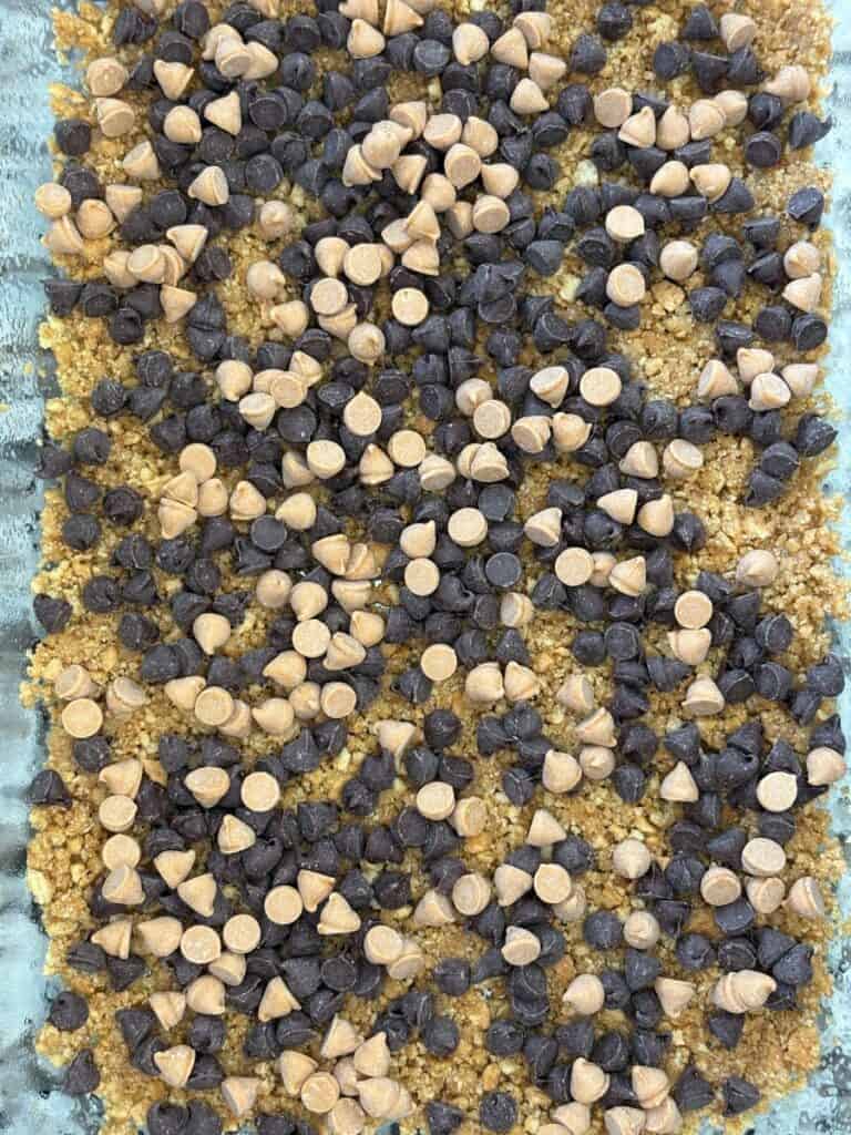 A close-up of a dessert base topped with a mix of chocolate chips and butterscotch chips, spread evenly over a crumbly, golden crust in a baking dish.