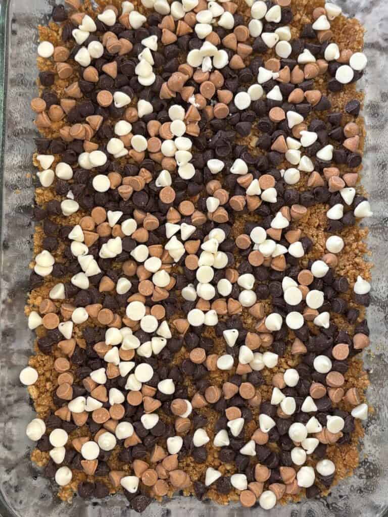 A close-up of a dessert in a glass baking dish, topped with a layer of chocolate, white, and butterscotch chips over a crumbly brown base.