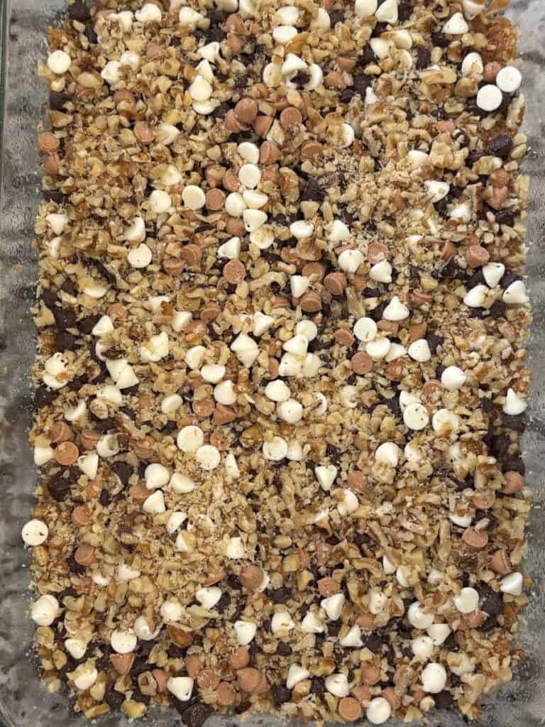 A baking dish filled with a layered dessert topped with chopped nuts, white and dark chocolate chips, and butterscotch chips, creating a textured and colorful surface.