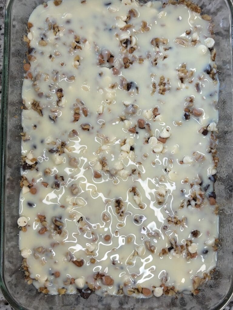 A glass baking dish filled with a layered dessert topped with sweetened condensed milk, nuts, and chocolate chips, ready to be baked.