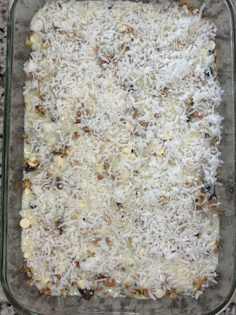 A glass baking dish filled with a dessert topped with shredded coconut, chopped nuts, and white chocolate chips, over a creamy, slightly golden layer.
