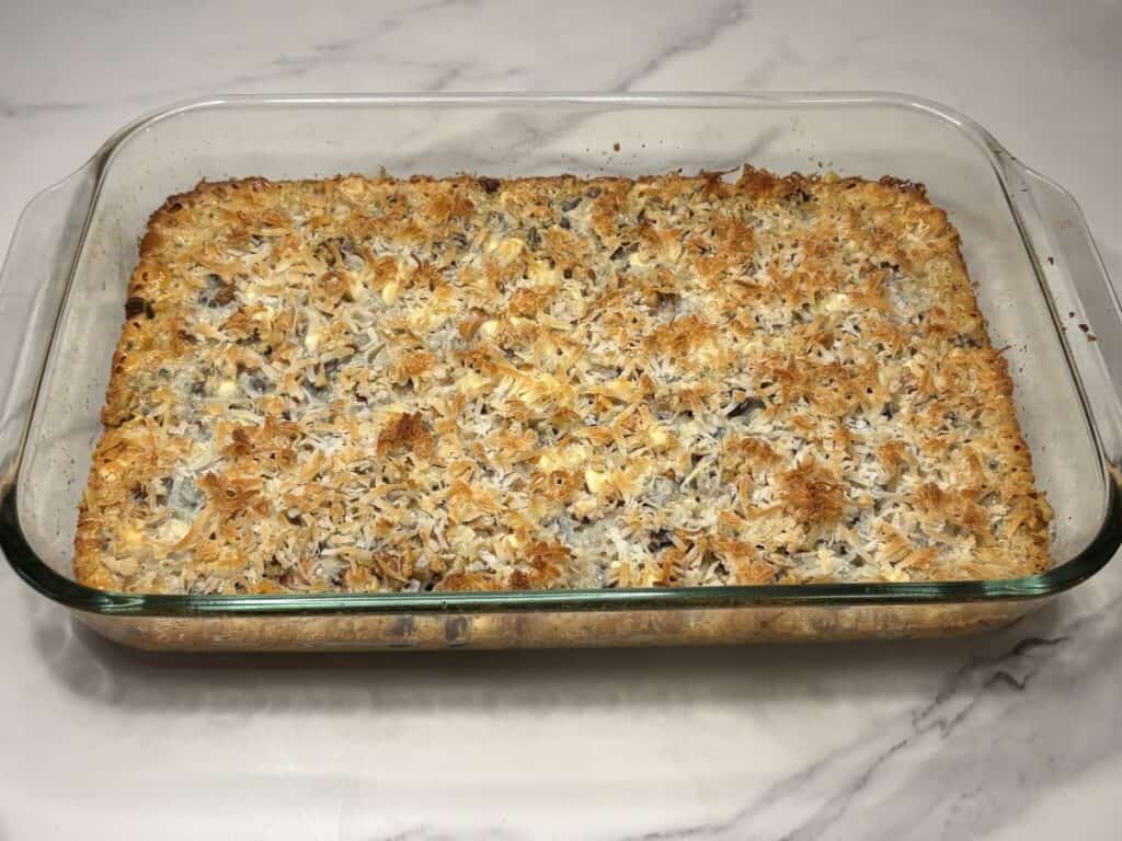 A glass baking dish filled with a golden-brown, baked dessert topped with toasted shredded coconut, resting on a light marble surface.