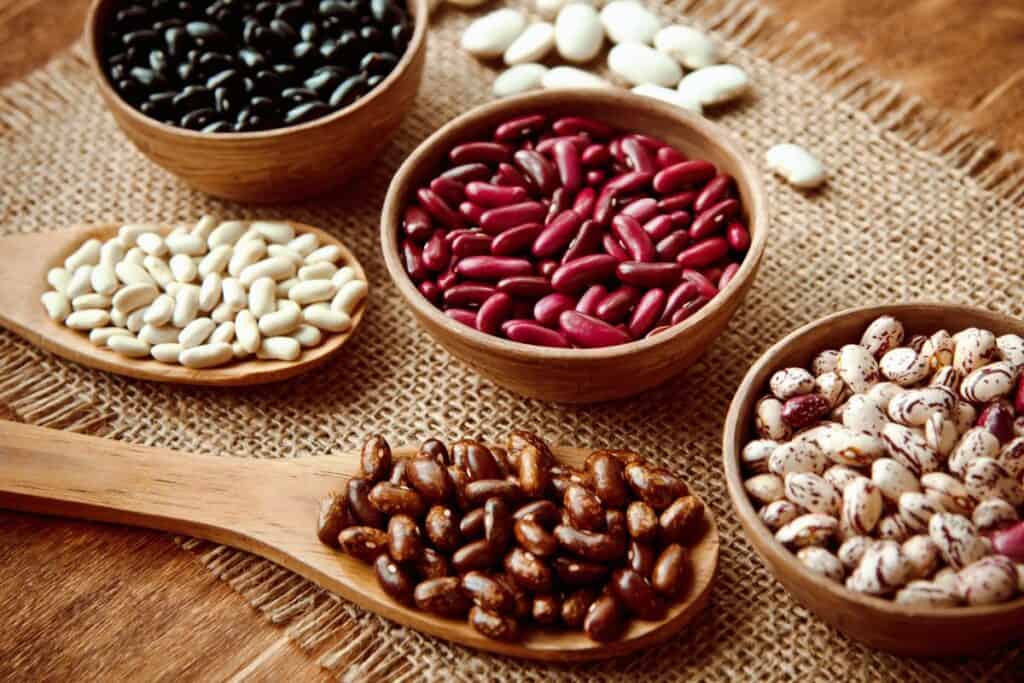 Five types of dried beans in wooden bowls and a spoon, arranged on a piece of burlap on a wooden table. The beans are various colors, including black, red, white, brown, and speckled.