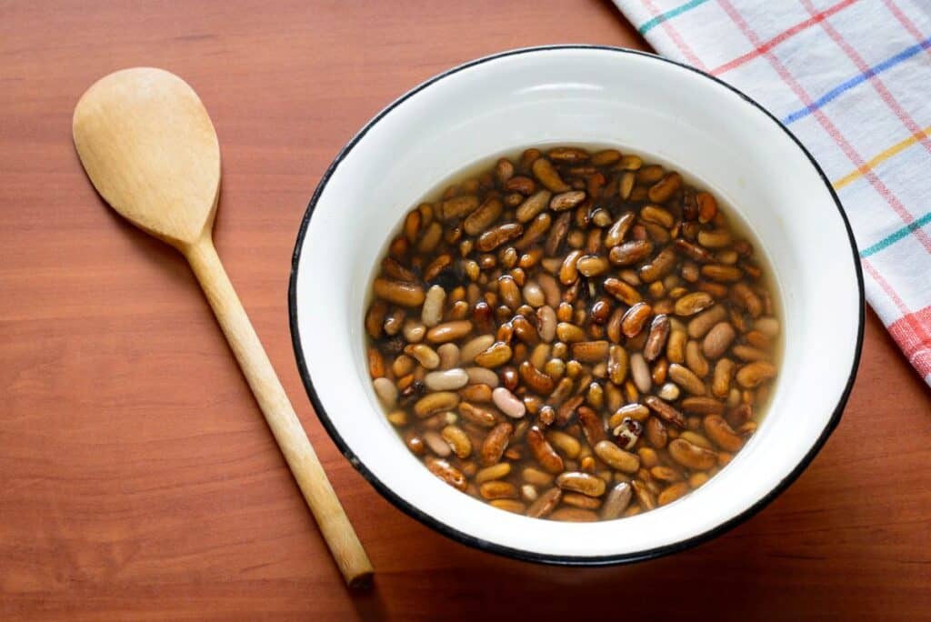 A white bowl filled with beans soaking in water sits on a wooden surface next to a wooden spoon, with a colorful checkered cloth in the background.