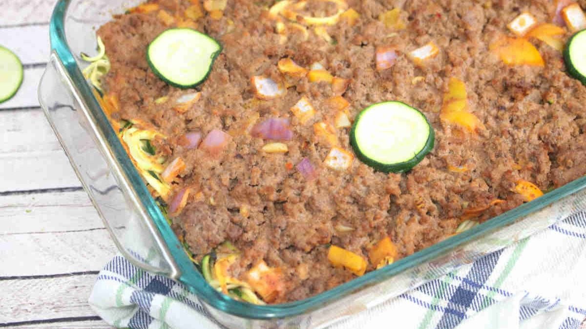 A glass baking dish filled with a casserole topped with browned ground meat, chopped yellow peppers, red onions, and several slices of zucchini, resting on a plaid cloth on a wooden surface.