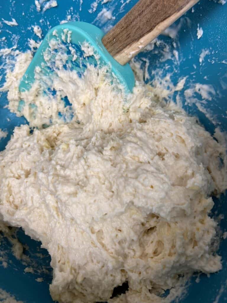 A close-up of thick, sticky bread dough being mixed in a blue bowl with a teal spatula and wooden handle. The dough has a rough, lumpy texture and appears partially combined.