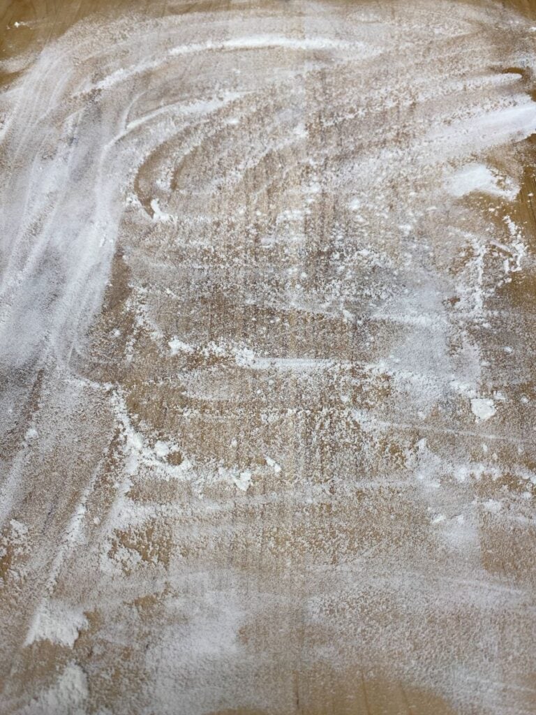 A wooden surface lightly dusted with scattered flour, showing swirled and uneven patterns left by brushing or spreading the flour.