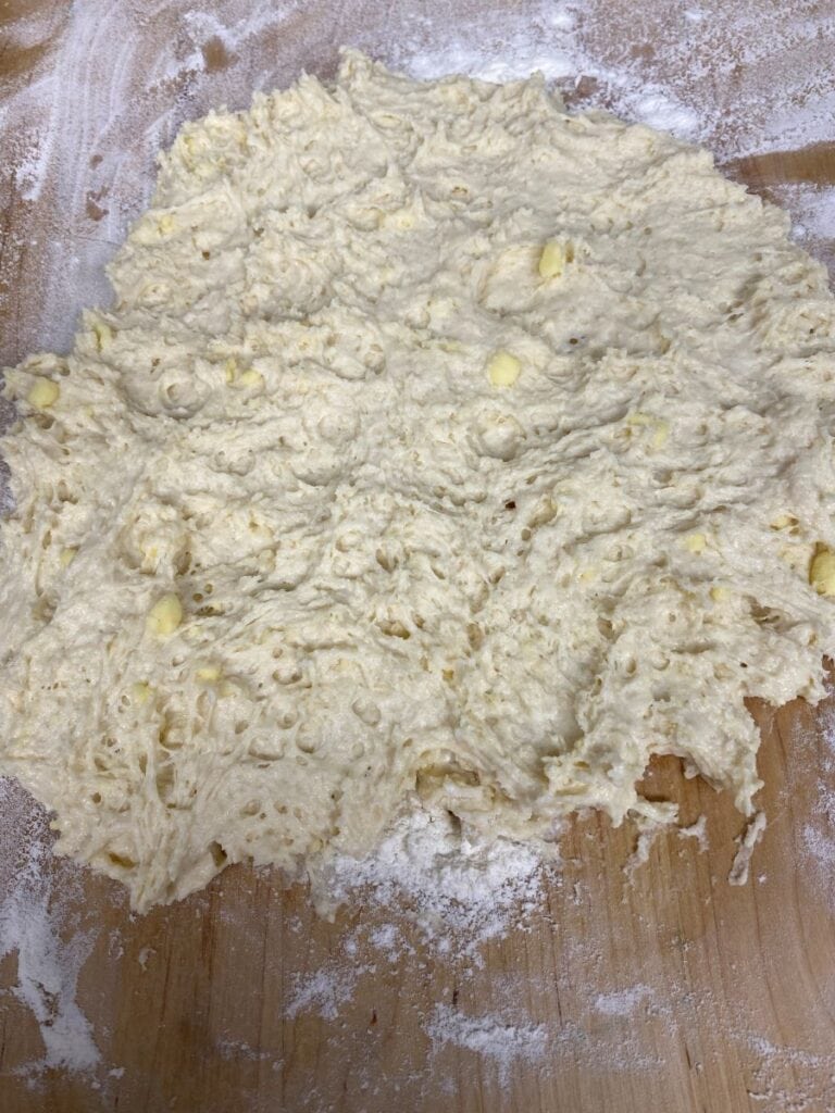 A rough, sticky dough with visible air bubbles and small chunks, spread out on a flour-dusted wooden surface, ready to be kneaded or shaped.