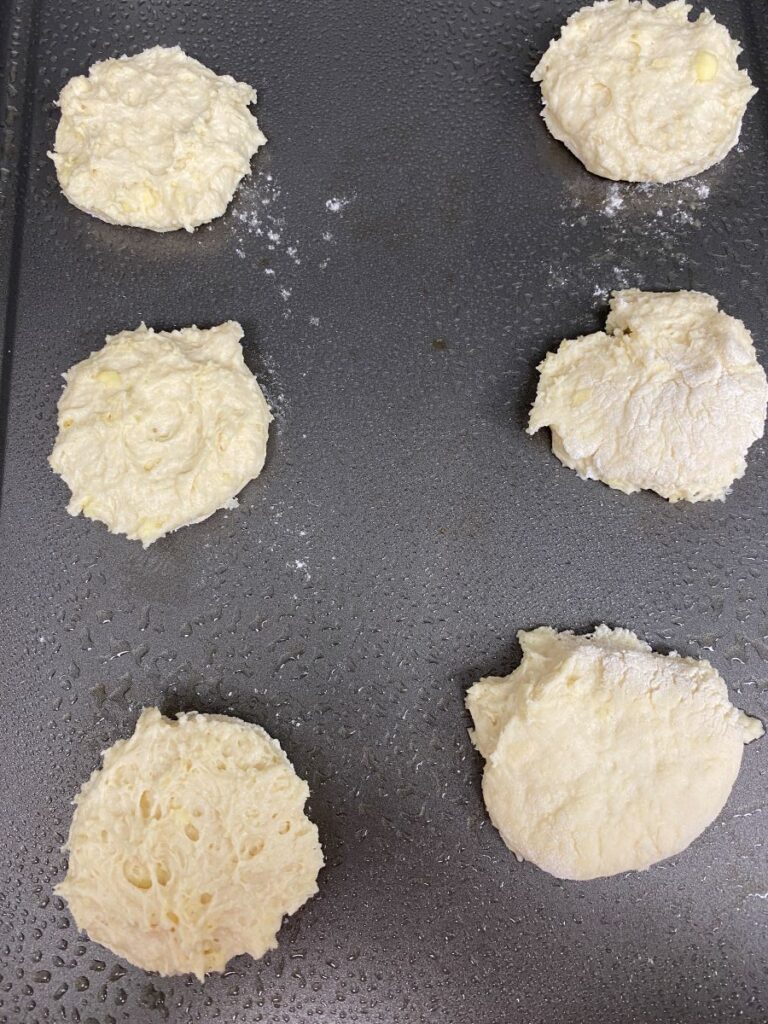 Six unbaked biscuit dough rounds are spaced out on a greased baking sheet, ready to go into the oven. The dough appears soft and slightly sticky with a rough texture.