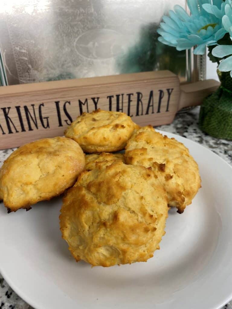 Four golden-brown biscuits on a white plate, with a rolling pin in the background labeled BAKING IS MY THERAPY and blue artificial flowers beside it.