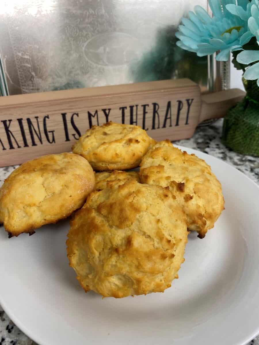 Four golden-brown biscuits on a white plate, with a rolling pin in the background labeled BAKING IS MY THERAPY and blue artificial flowers beside it.
