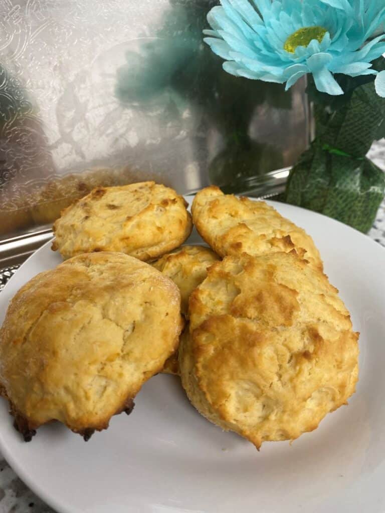 A white plate with five golden-brown biscuits sits on a counter, next to a turquoise artificial flower in a green vase, with a silver tray in the background.
