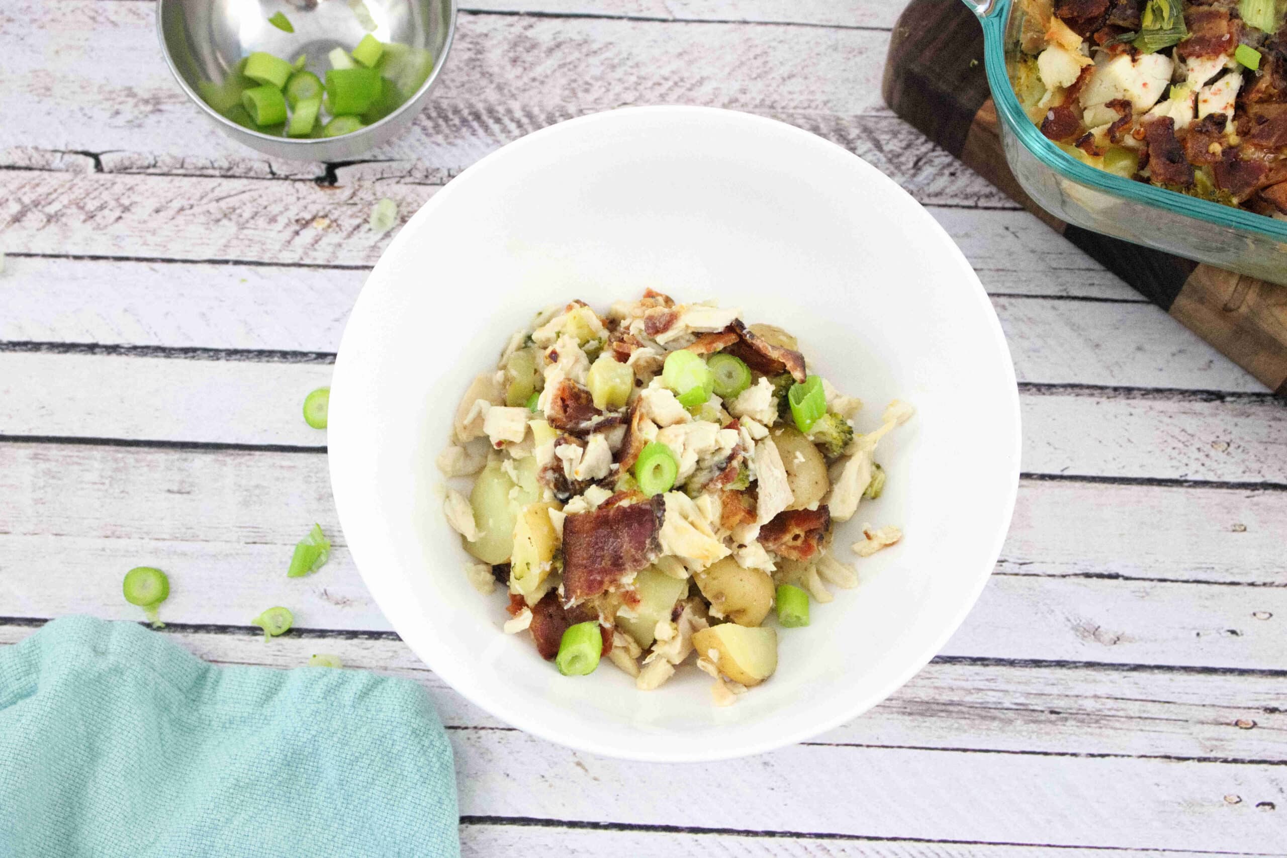 A white bowl filled with a salad made of chopped potatoes, chicken, bacon, and sliced green onions sits on a rustic white wooden surface near a blue cloth. A baking dish and a small bowl of green onions are nearby.