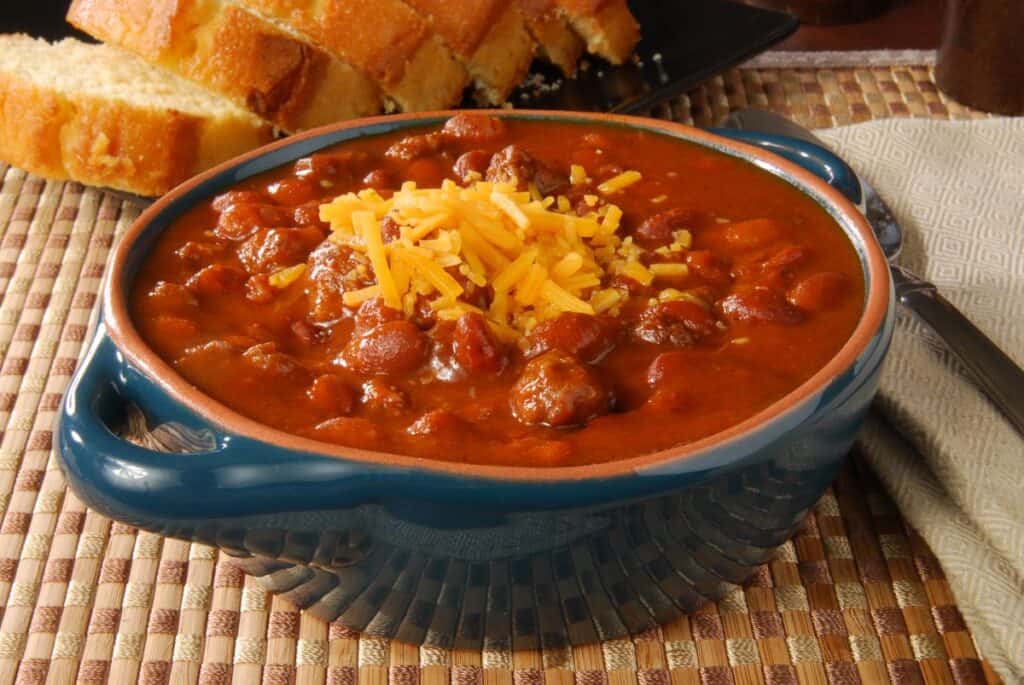 A bowl of chili topped with shredded cheddar cheese sits on a woven placemat, with slices of cornbread on a plate in the background and a fork resting nearby.