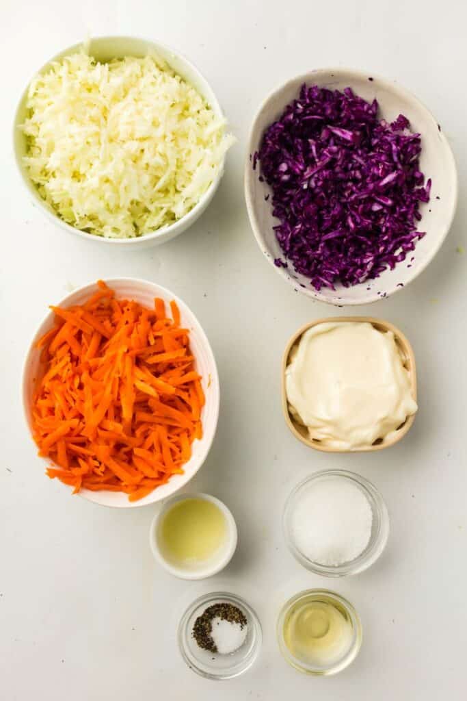Bowls of shredded green cabbage, purple cabbage, and carrots, along with small bowls of mayonnaise, vinegar, sugar, salt, and pepper, are arranged on a white surface, ready for making coleslaw.